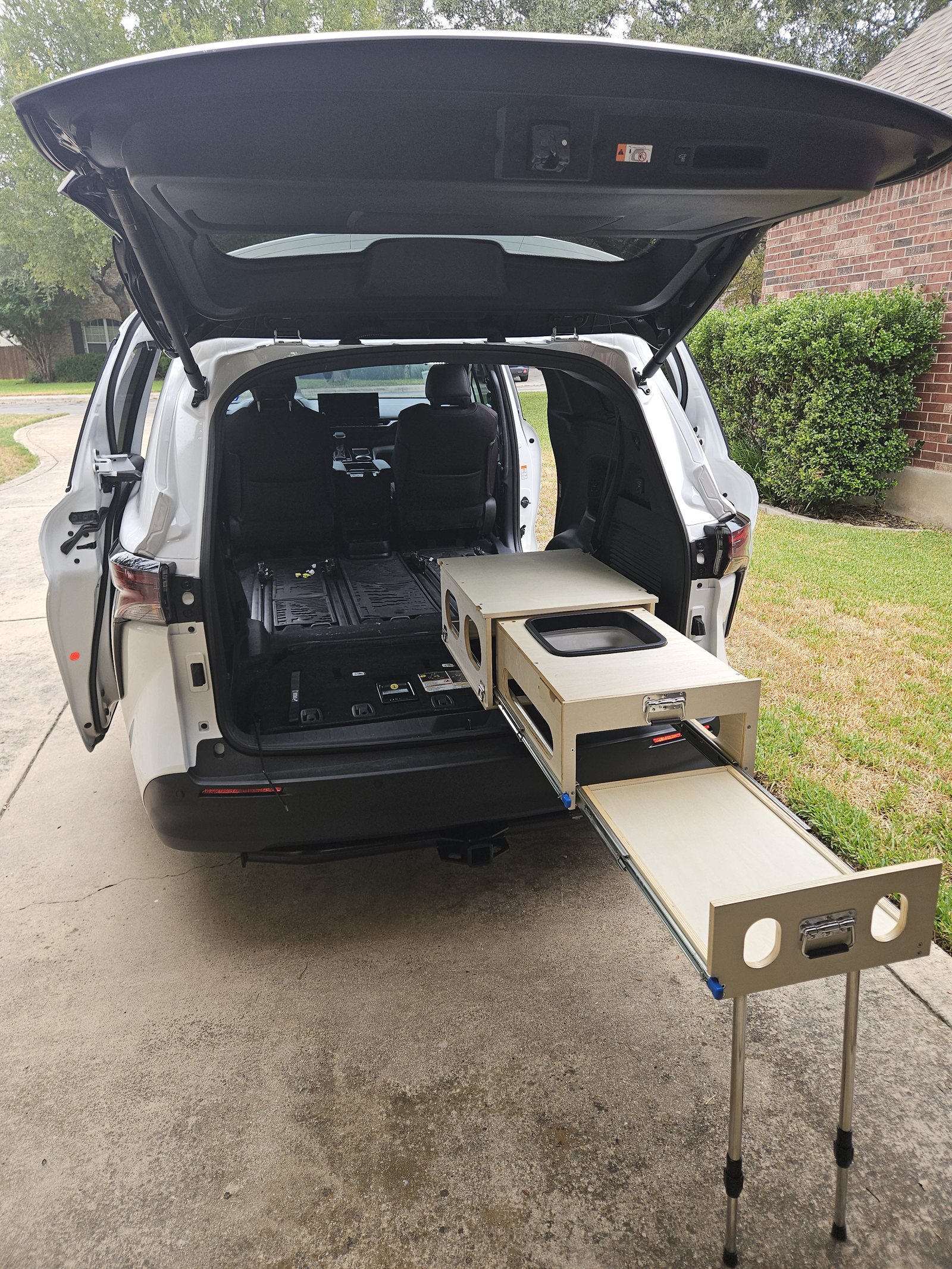 The slide-out camp kitchen pulled out of the open tailgate of a white Sienna on a driveway, worktop folded out on its legs, with the second-row floor empty behind it
