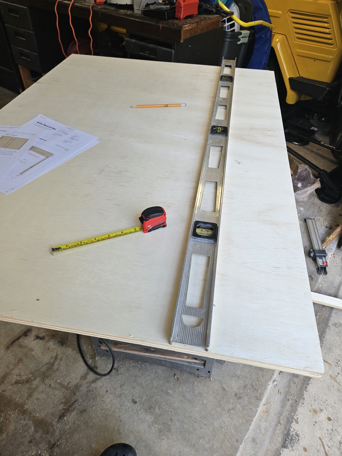 A large plywood panel on the saw table with the printed cut plan, a four-foot level, a tape measure and a pencil laid on it